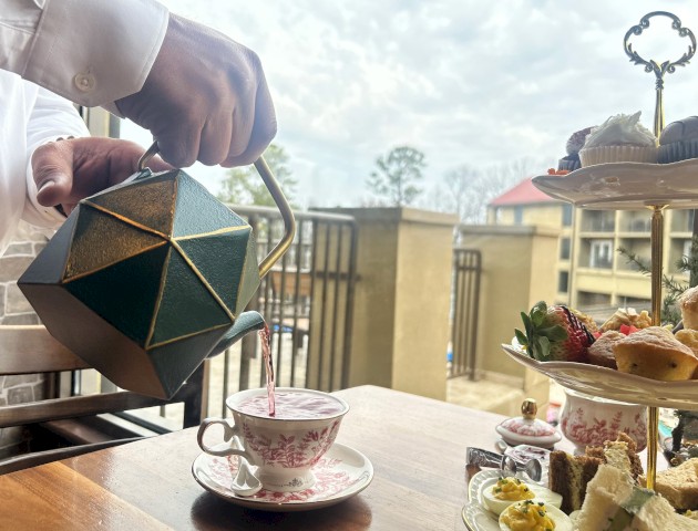 A hand pouring tea from a geometric teapot into a teacup on a table beside a tiered pastry stand and a scenic outdoor view, afternoon light.