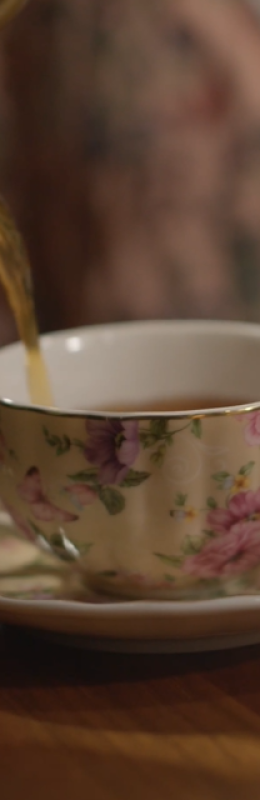 A teapot pours hot tea into a delicate floral teacup set on a wooden table, with a saucer, spoon, and plate in the background.