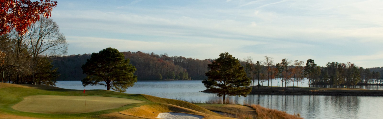 A serene golf course by a calm lake, with sand bunkers, green fairways, and trees reflecting in the water, under a clear blue sky.