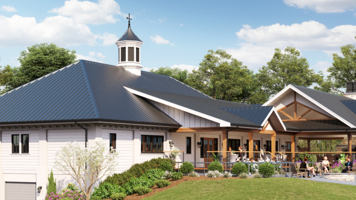 A single-story clubhouse with a blue metal roof, cupola, large glass entry, surrounding patio, and a green lawn under a bright, partly cloudy sky.