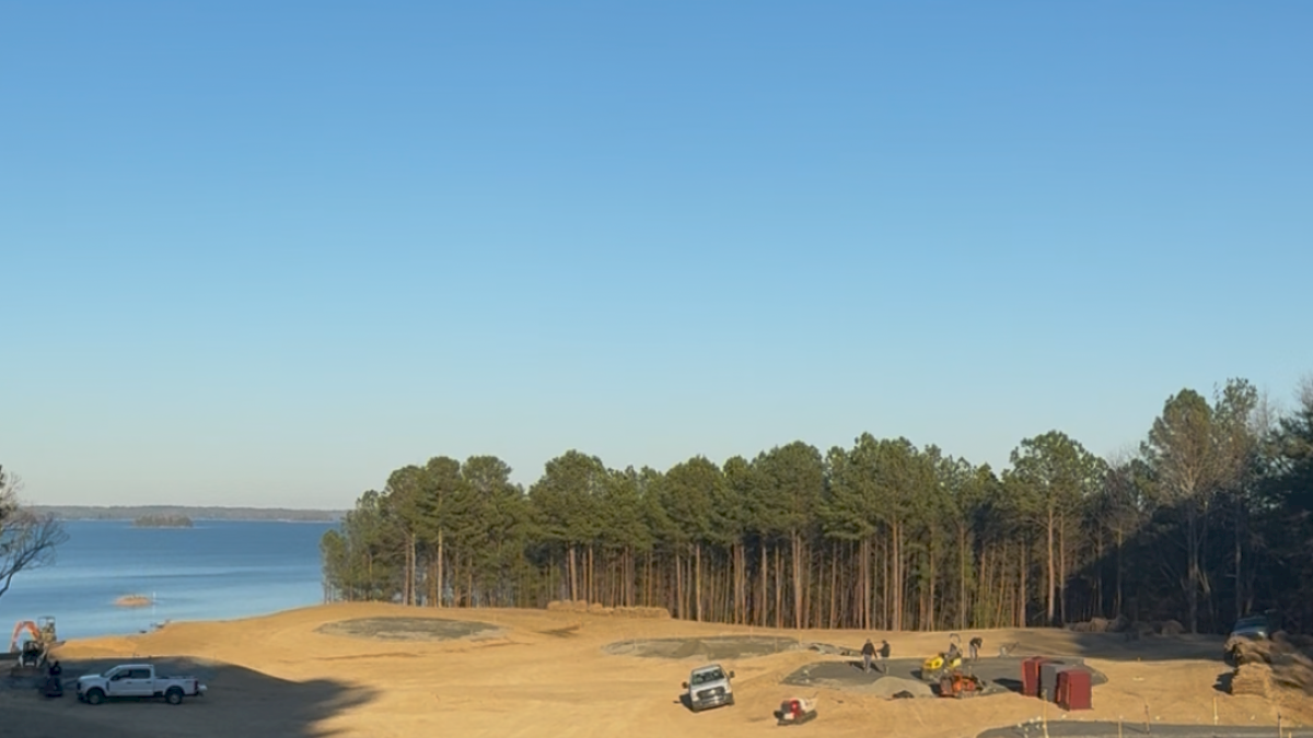 A sandy lakeshore beach with a line of trees in the distance, a few parked cars and tents, and calm blue sky above. End.