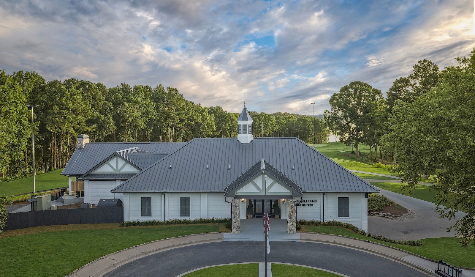 A golf clubhouse with a circular driveway, surrounded by green trees and a partly cloudy sky above.