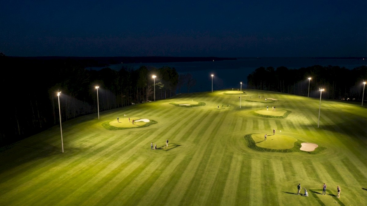 Nighttime view of a lit golf practice area with several sand bunkers, neatly mowed green grass, and a few people practicing.