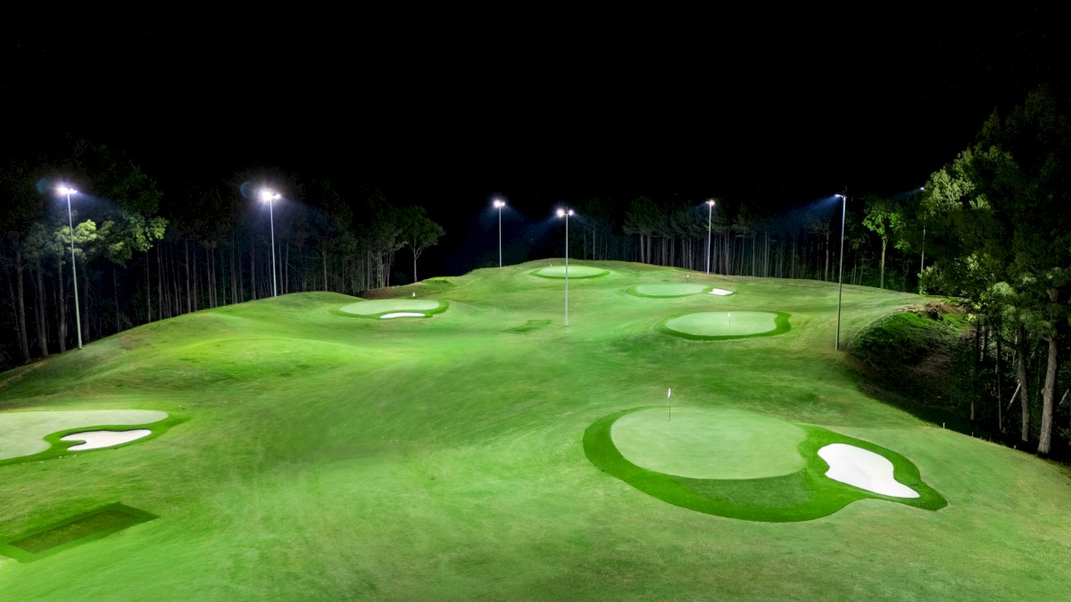 A golf course at night, lit by tall lights, with multiple putting greens and surrounding trees.