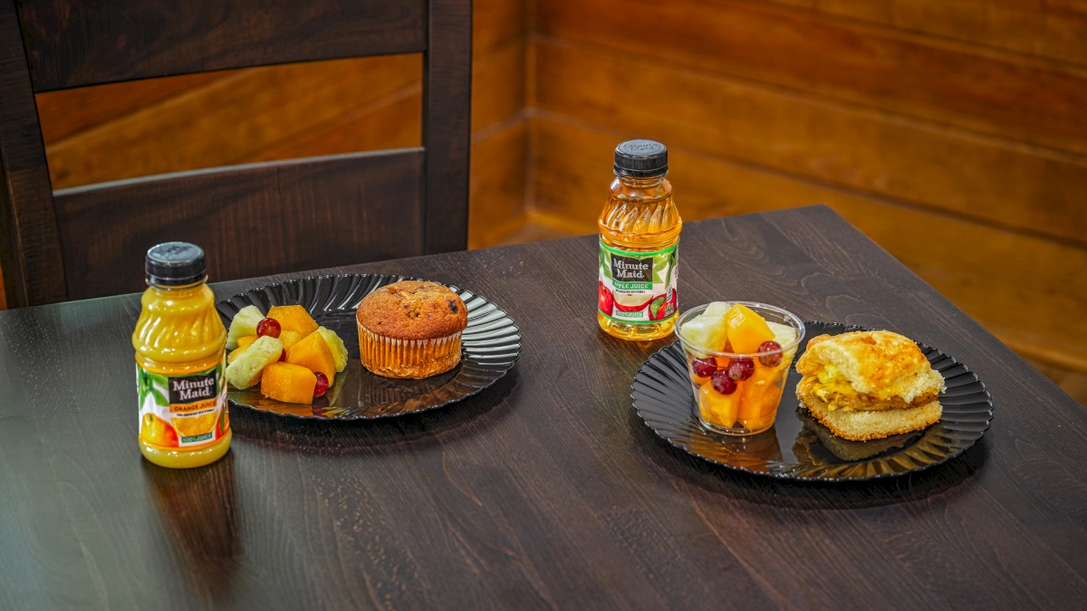 A table with two plates of fruit, muffin, and a sandwich, along with two bottles of orange juice on a dark wooden surface.