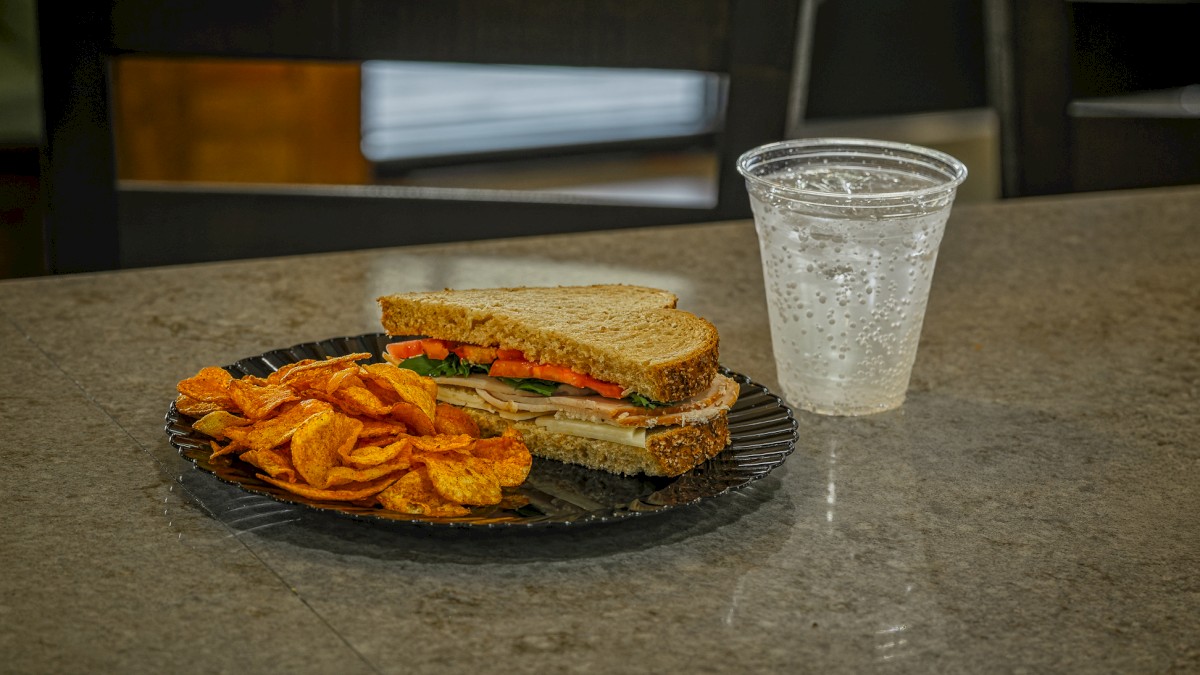 A turkey, ham, and veggie sandwich with potato chips and a soda on a cafeteria table.