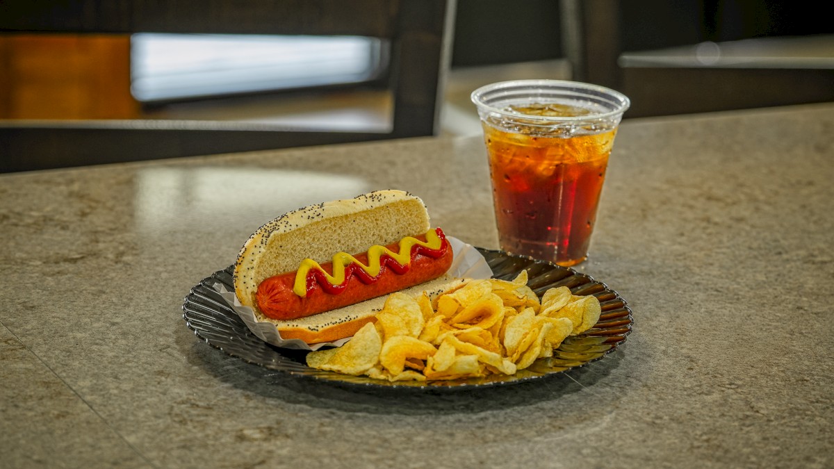 A hot dog with mustard and ketchup, potato chips, and a cup of iced soda on a restaurant counter.