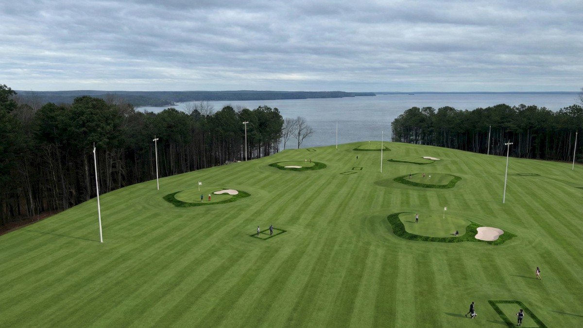 A grassy golf practice range with multiple green targets, sand bunkers, light poles, and a distant ocean view under a cloudy sky.