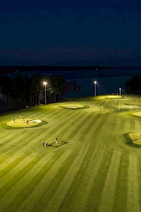 A nighttime golf practice field with several sand bunkers, bright floodlights, and a few people near the greens, illuminated long into the dark.