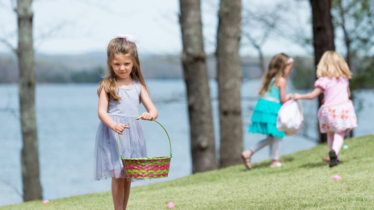 Three little girls in a park by the water; one in the foreground holding a basket, two others walking away.