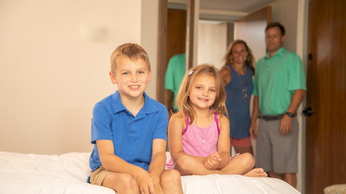 Two kids sit on a bed smiling at the camera, with two adults in the doorway behind them in a cozy home setting.