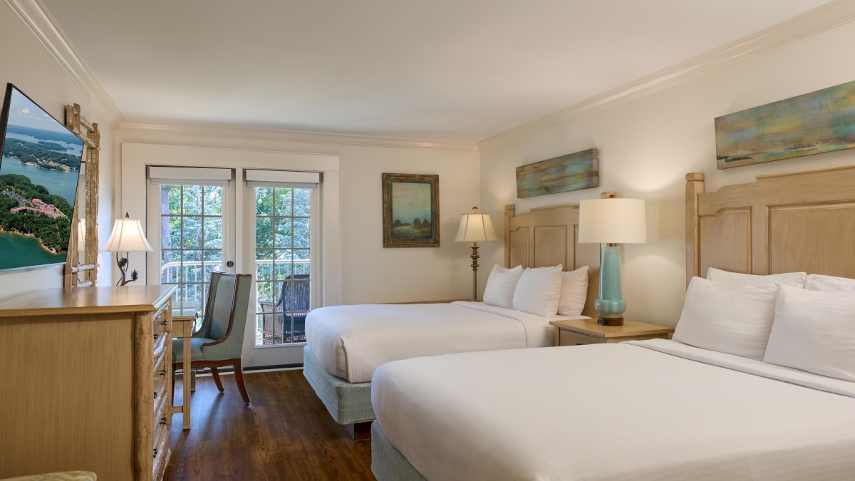 A bright hotel room with two queen beds, a desk and chair, lamps, artwork, and a balcony door overlooking greenery.