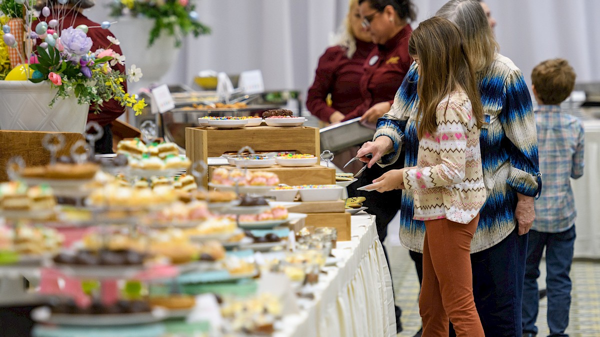 A family-friendly bake sale or buffet with a long table of assorted pastries and treats; kids browse and adults assist, colorful display.