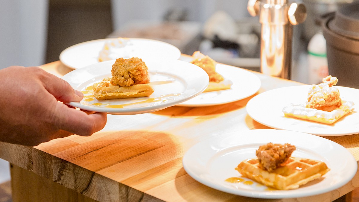 A chef plates three small dishes with fried topping on crispy bases, served on white plates along a wooden counter in a kitchen.