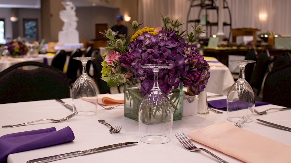 A formal banquet table with a purple floral centerpiece, neatly arranged silverware, napkins, and glassware in a decorated event hall.
