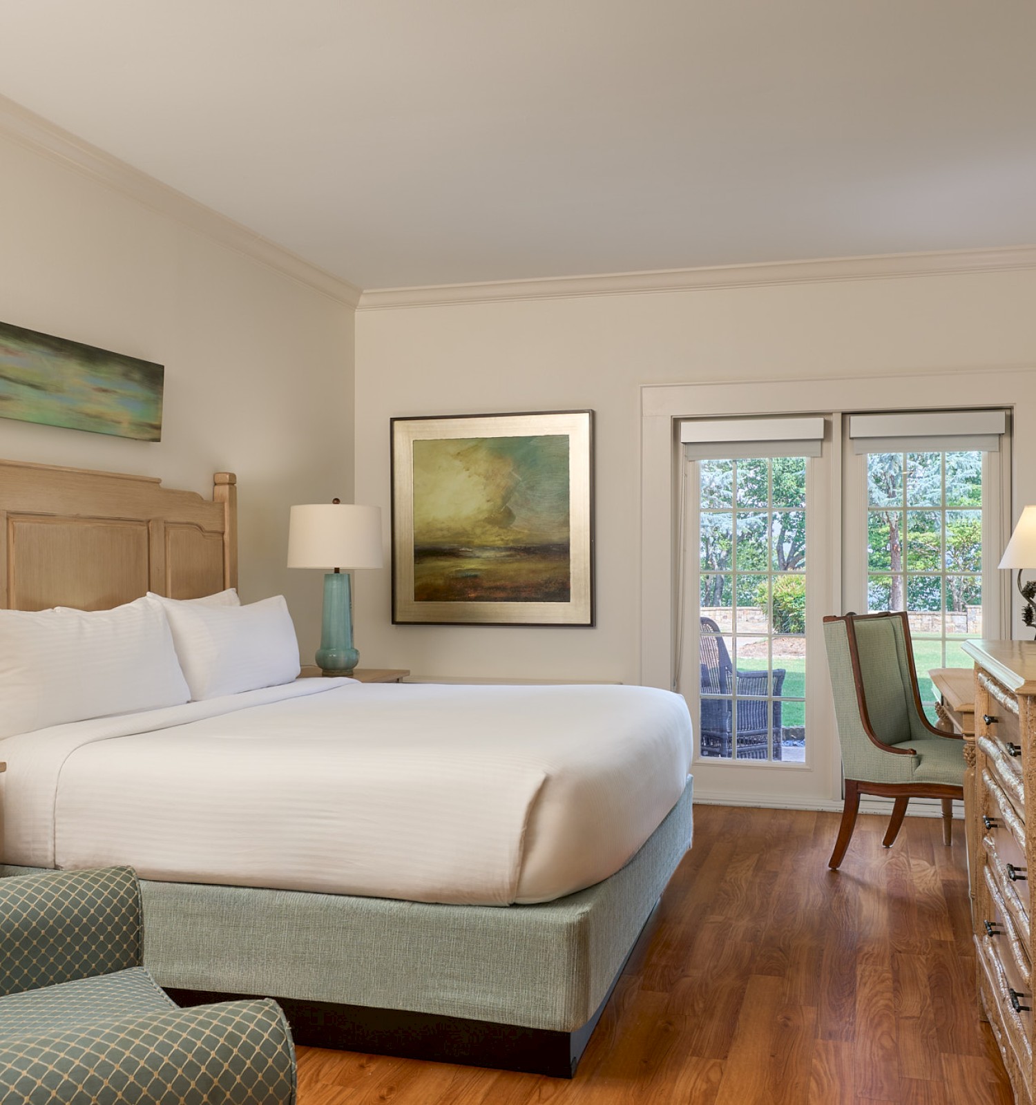 A cozy hotel bedroom with a large bed, artwork, lamps, a dresser, and patio access, overlooking a lush green landscape and water.