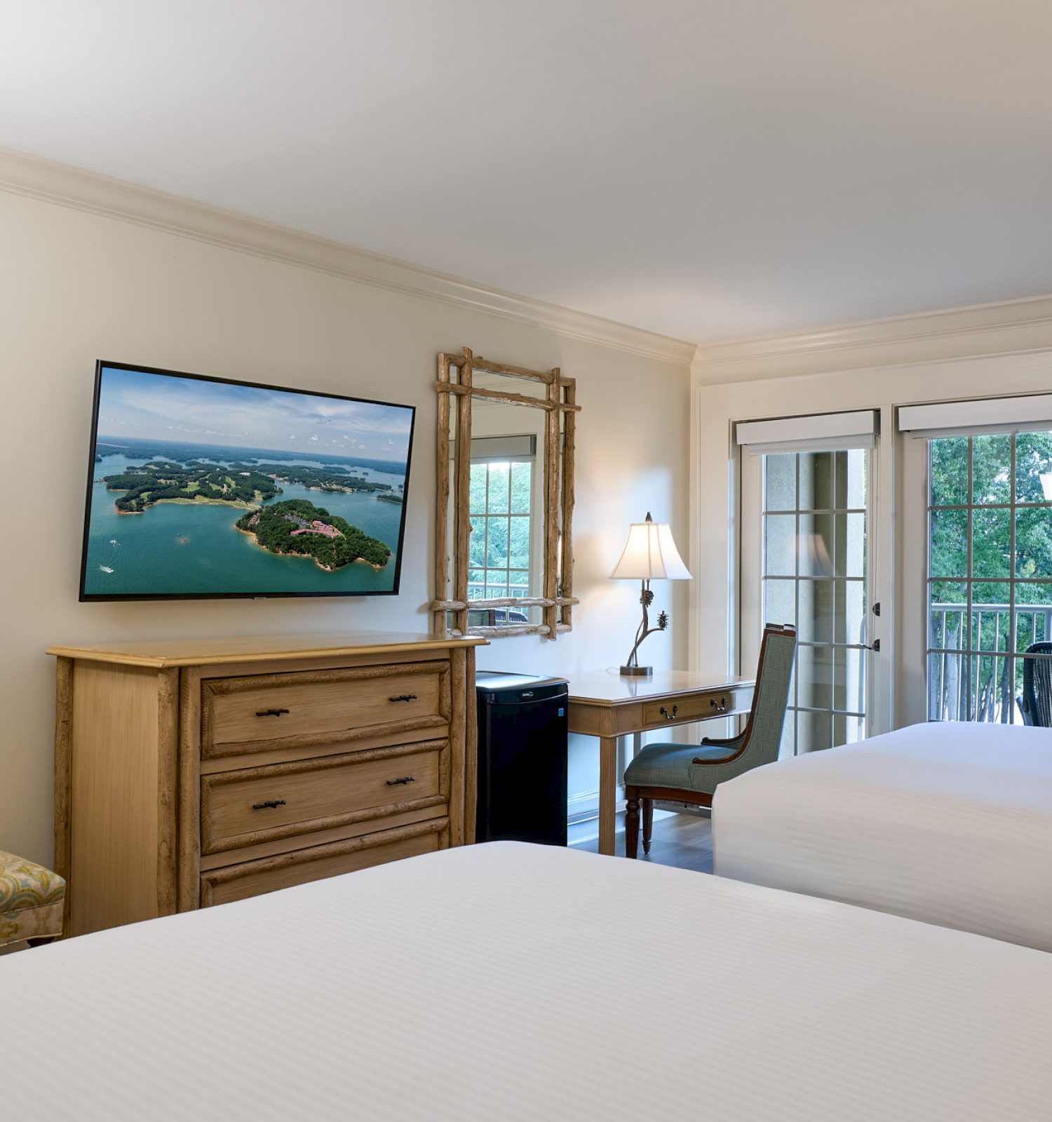 A cozy hotel room with two beds, a TV, a writing desk, a chair, artwork, and a balcony door with outdoor greenery.