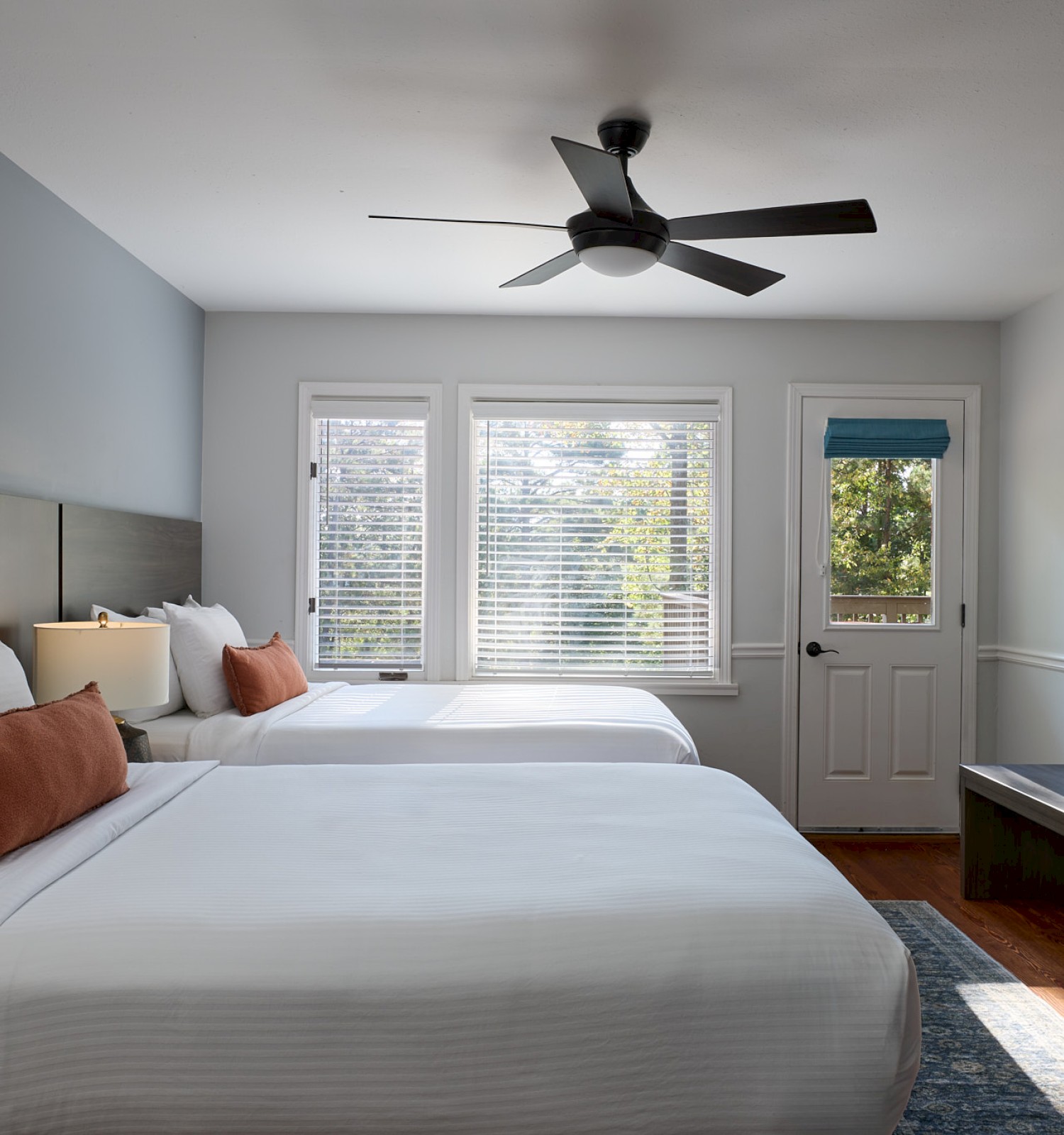 A bright, modern bedroom features two white beds with rust-colored pillows, a wall-mounted TV, and large windows showing greenery outside.