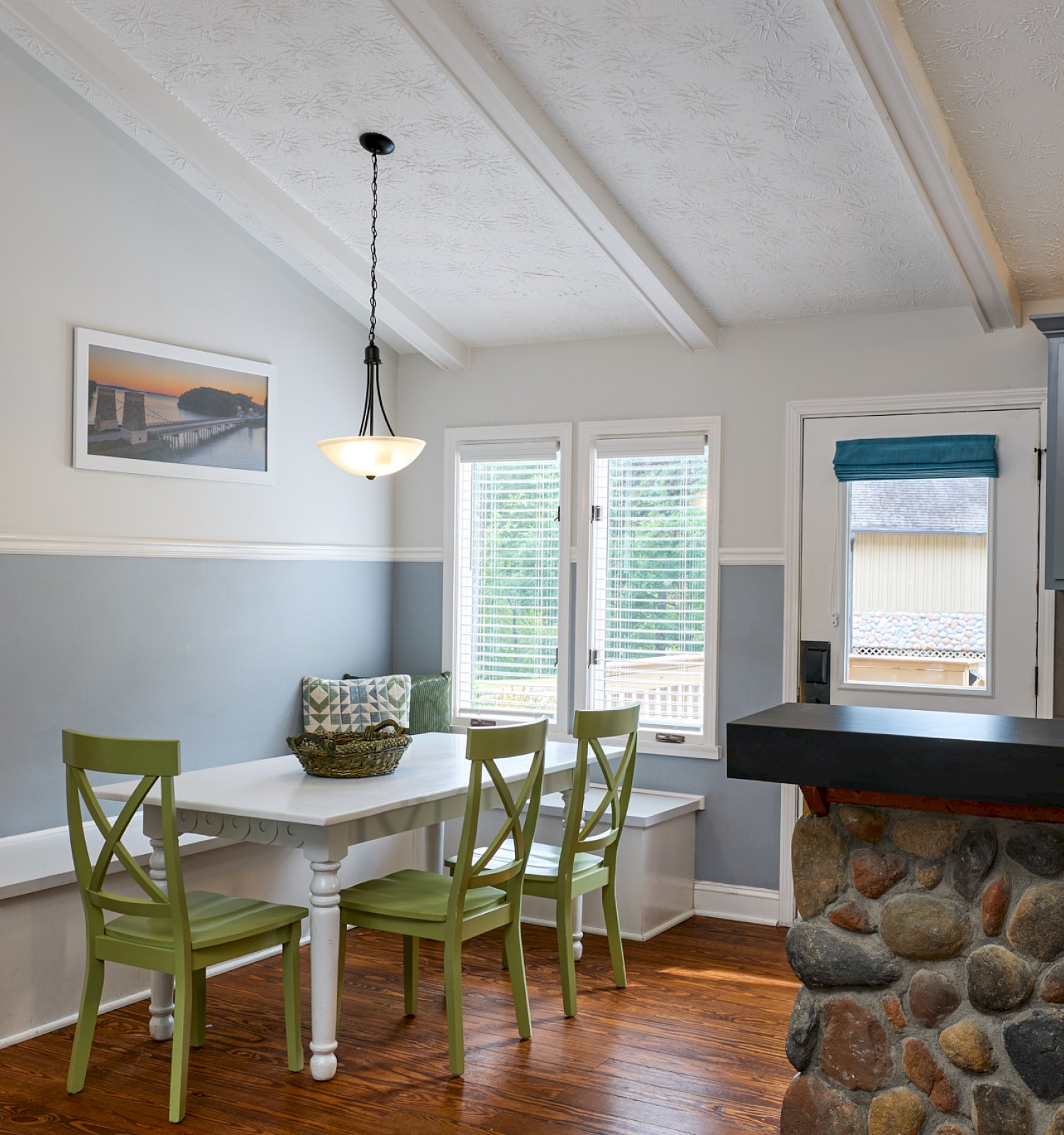 A cozy kitchen with a dining area featuring a white table, green chairs, and a stone kitchen island, illuminated by natural light.
