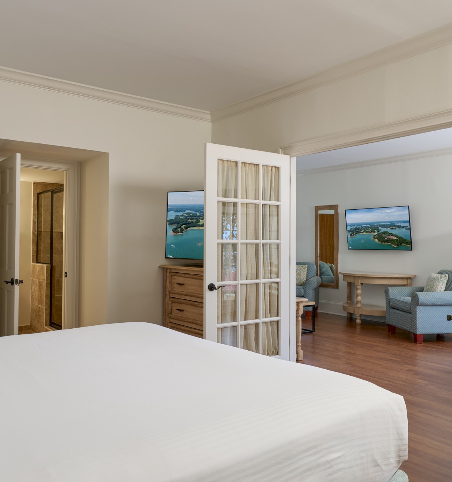 A bedroom with a view into a sitting area, featuring a bed, TV, armchairs, and a doorway to a bathroom or closet space.