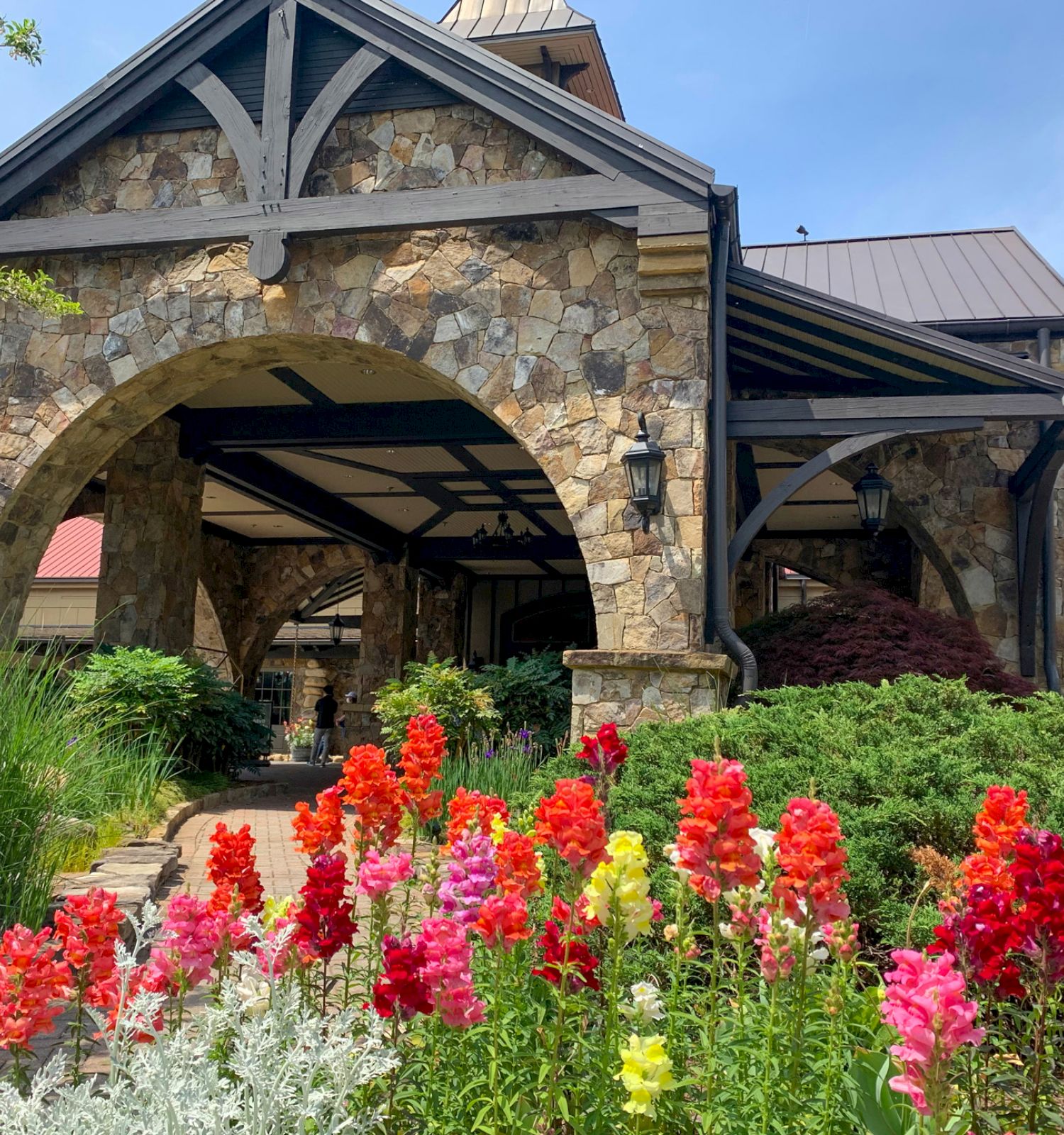 A stone entrance with a wooden roof, leading to a building, amidst colorful blooming flowers under a blue sky.