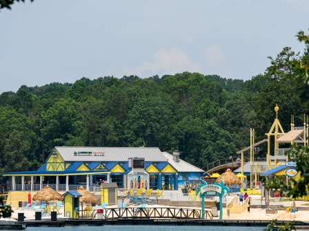 A lakeside amusement area with colorful buildings, slides, and a dock; bright blues and yellows, trees in the background, calm water—fun family fun.