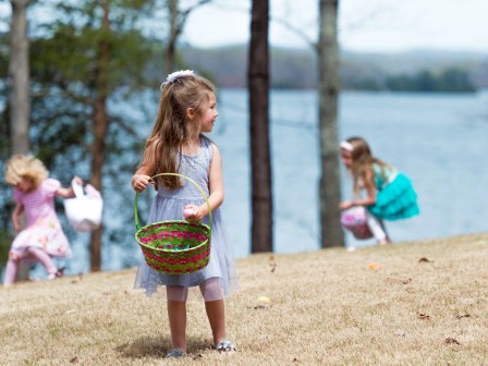 Three kids hunt for Easter eggs on a grassy hill by a lake, with one girl in a gray dress holding a basket.