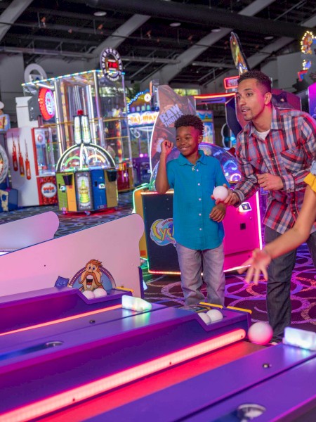 Children and an adult play skee-ball at an arcade with colorful neon lights and various games in the background.