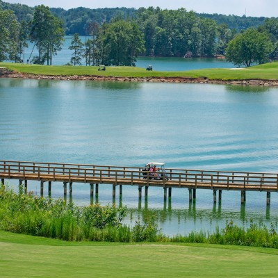 A golf course with a lake and a golf cart on a wooden bridge, surrounded by lush greenery and trees under a bright, sunny sky.