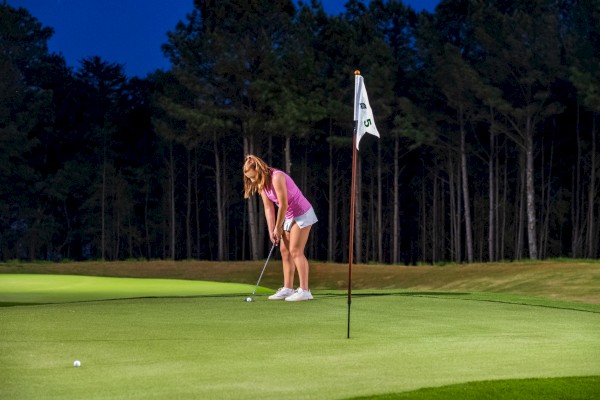 A woman is preparing to putt on a golf course during evening, surrounded by trees, under a darkening sky.