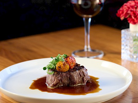 A gourmet steak topped with greens and sauce on a white plate, paired with a glass of red wine in a cozy setting.