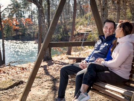 Two people sit on a wooden swing beside a lake in a forest, smiling and enjoying a sunny outdoor moment together.
