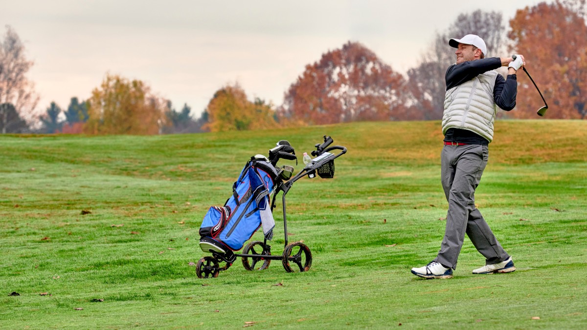 A golfer swings on a sunny course, with a blue golf bag on a push cart nearby; autumn trees in the background.