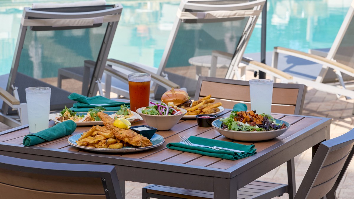 A dining table by a pool is set with salads, fried fish, French fries, a burger, drinks, and napkins on a sunny day.