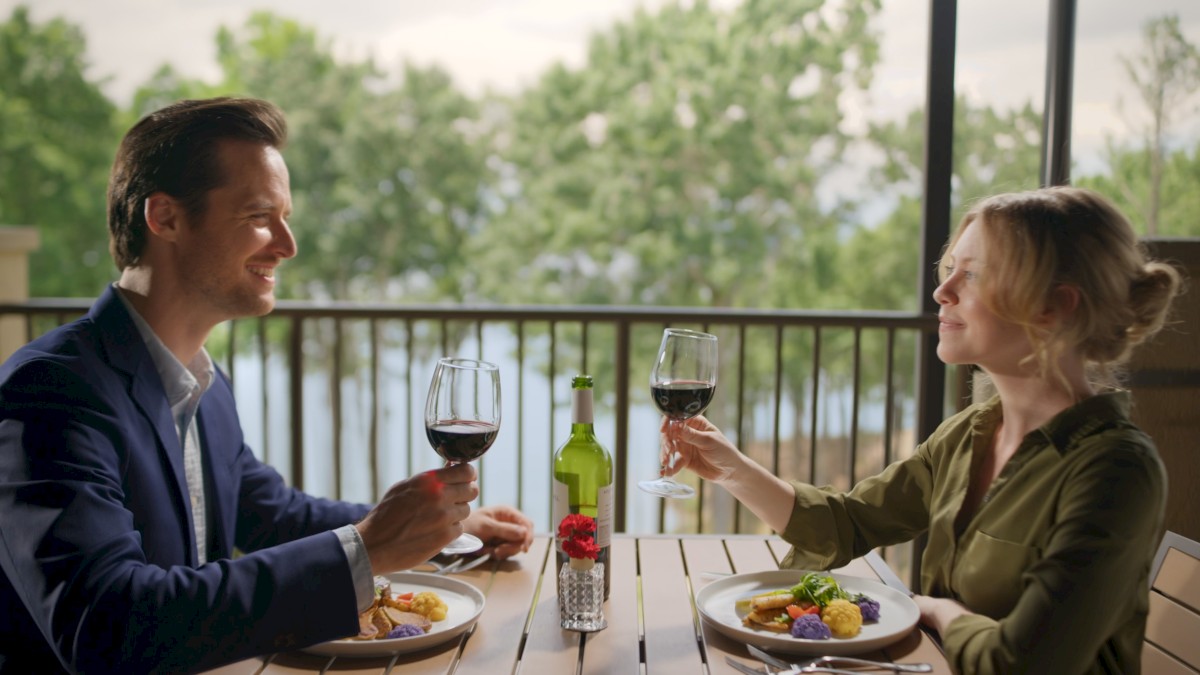 A man and woman share a toast with glasses of red wine during a meal on a balcony with a scenic view of trees and a water body.