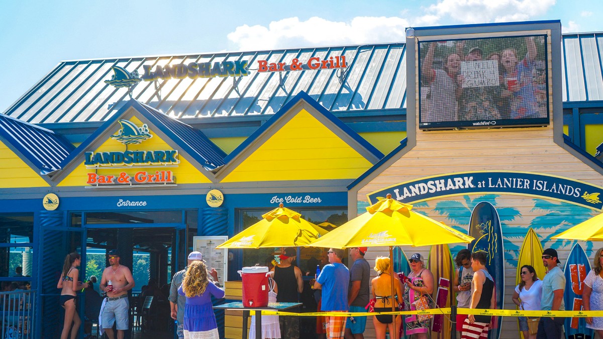 This image shows the entrance of LandShark Bar & Grill with people lined up outside under yellow umbrellas, featuring surfboards and a large screen above.