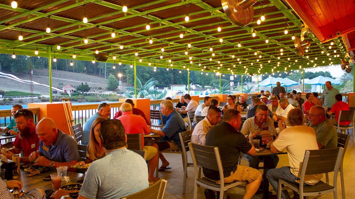 A group of people dining on an outdoor patio with string lights overhead, overlooking a scenic view with hills and a waterslide in the background.