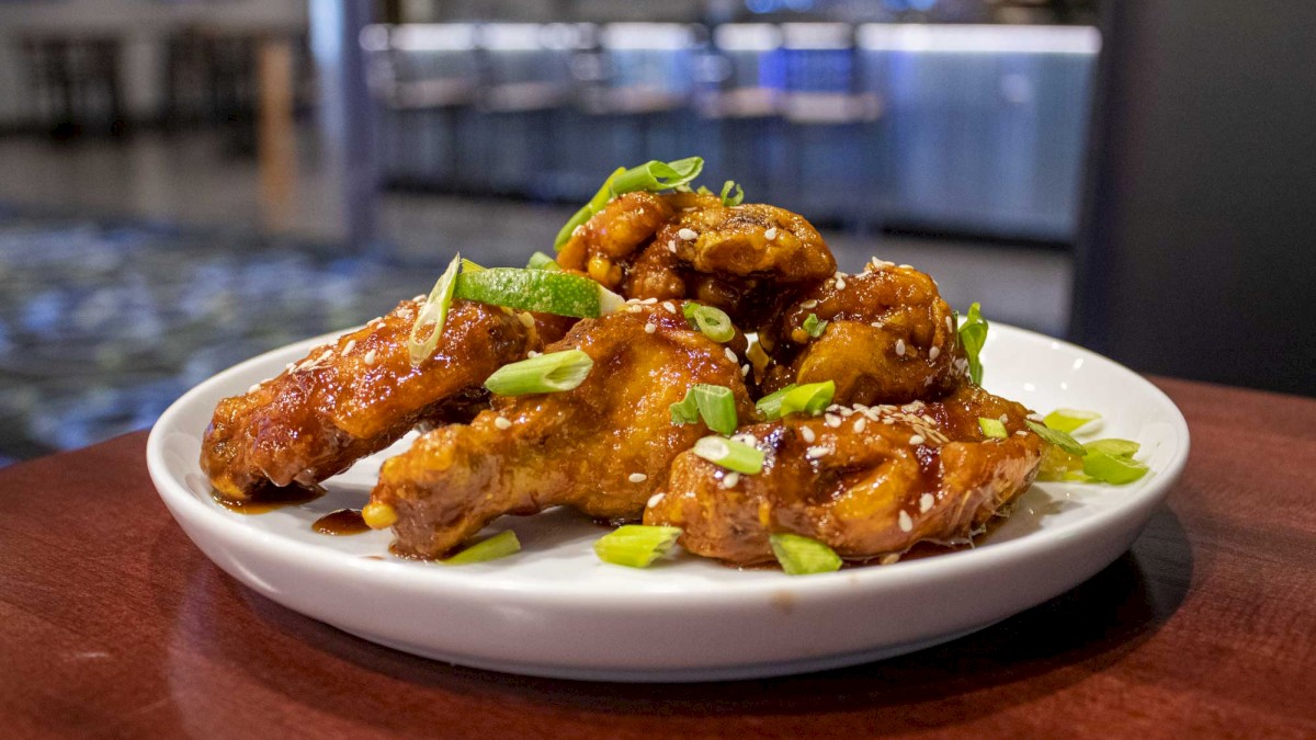 A plate of sauced chicken wings garnished with sesame seeds and chopped green onions, set on a wooden table in a restaurant setting.