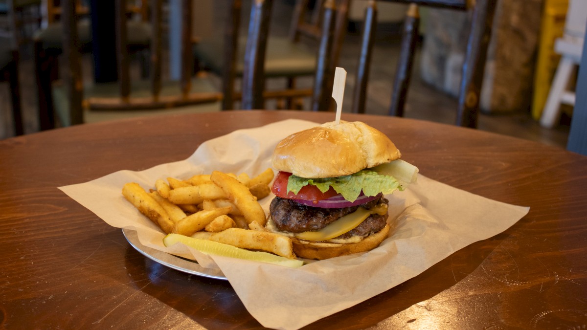 A cheeseburger with lettuce, tomato, and onion sits on a plate with fries on a wooden table. The table has rustic chairs around it.