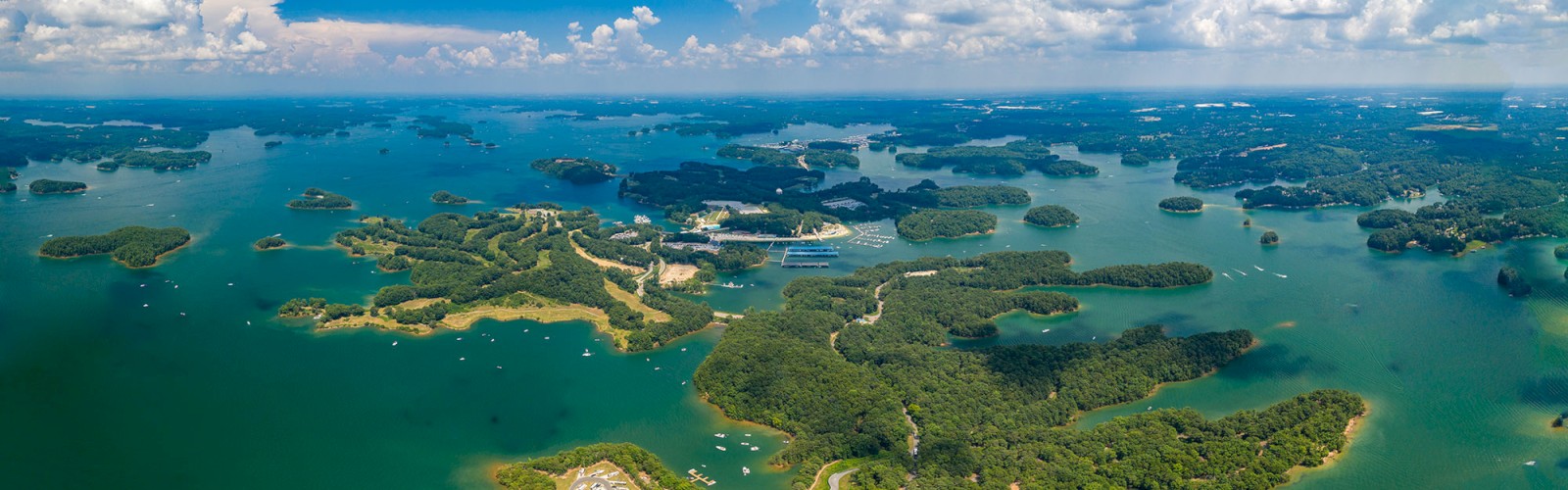 Aerial view of a lake with numerous tree-covered islands, boats, and a marina, under a blue sky with scattered clouds.