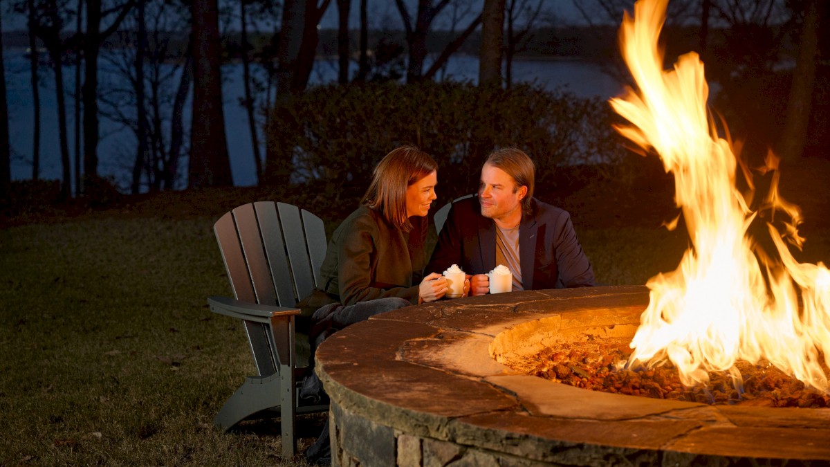 Two people sit by a lit outdoor fire pit, chatting warmly in the evening, a cozy moment under dark trees.