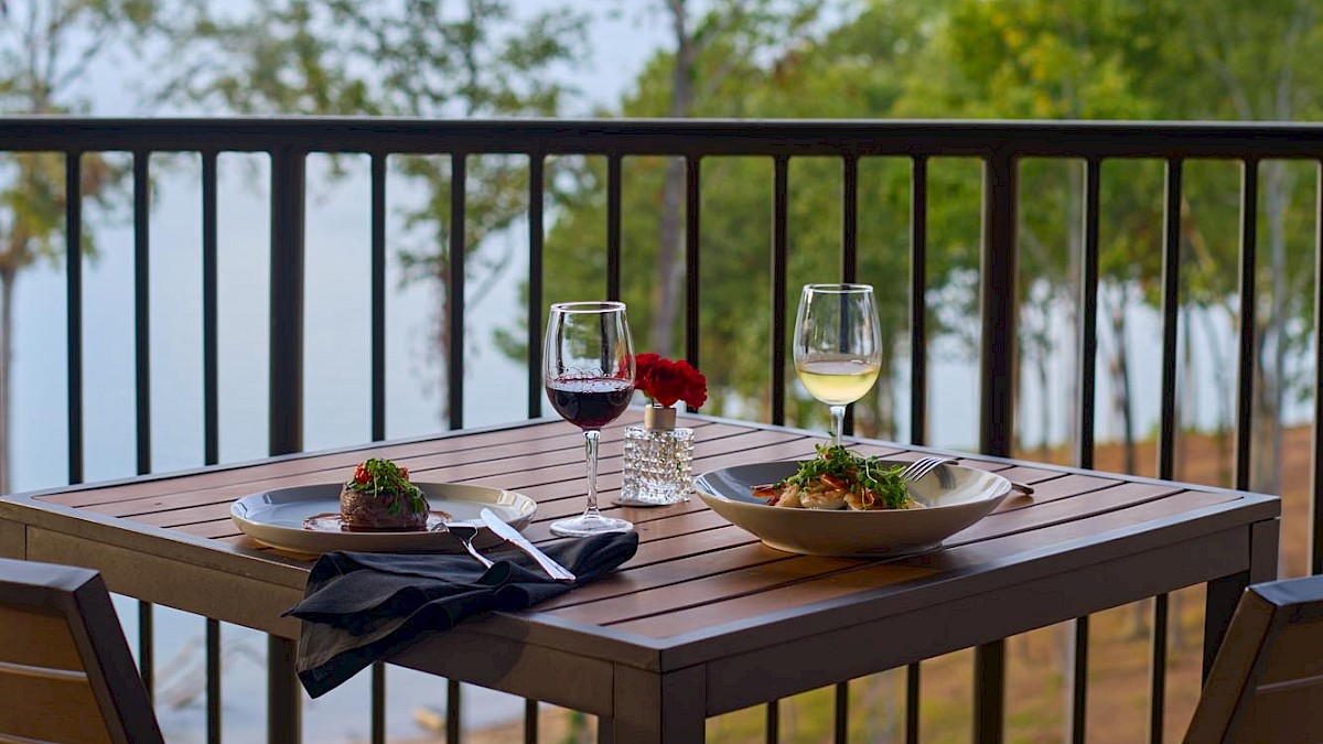A balcony table set for two with wine, two plates, and a dish; overlooking trees and a distant water view under a blue sky.