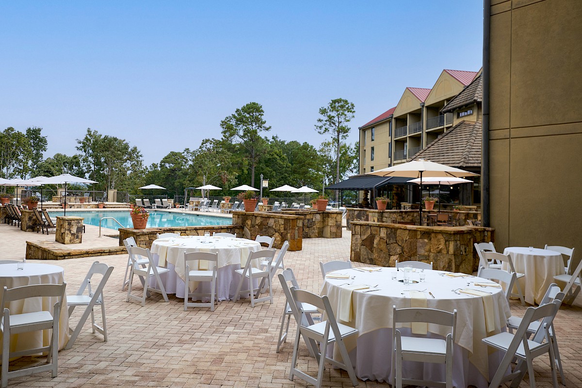 An outdoor poolside dining area with round tables, white chairs, umbrellas, and a pool in the background on a sunny day.