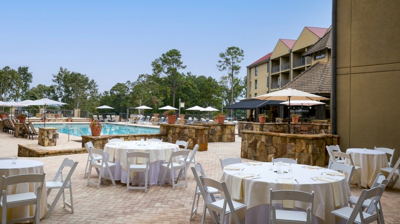 An outdoor poolside dining area with round tables, white chairs, umbrellas, and a pool in the background on a sunny day.