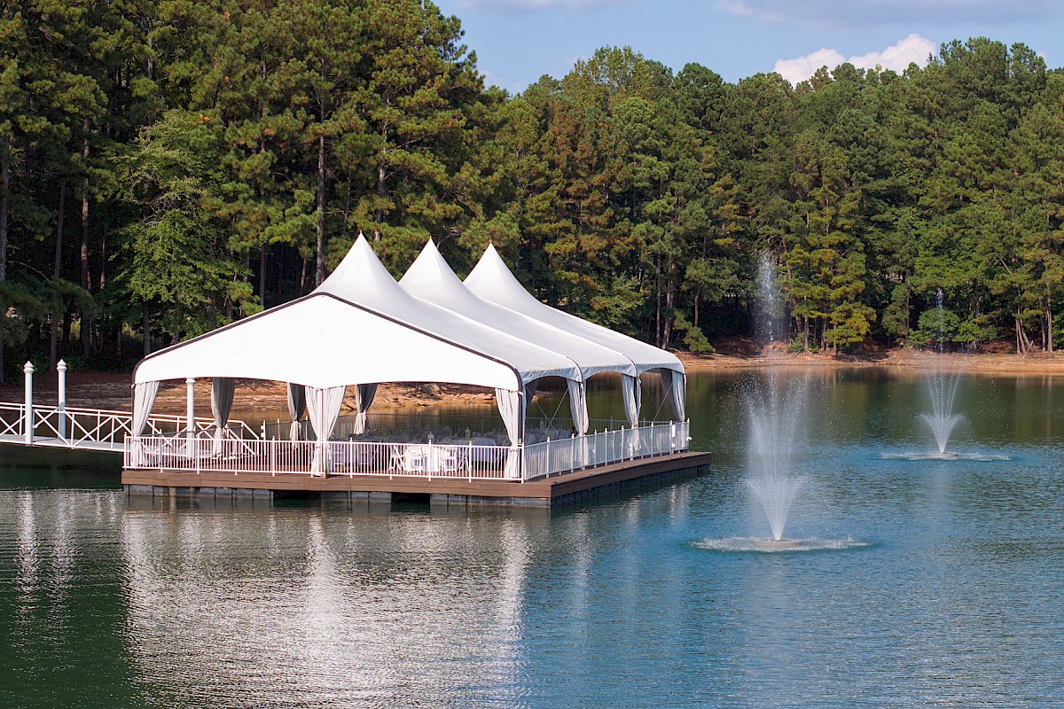 A white lakeside tent with fountains on a peaceful lake surrounded by a forested area under a partly cloudy sky.