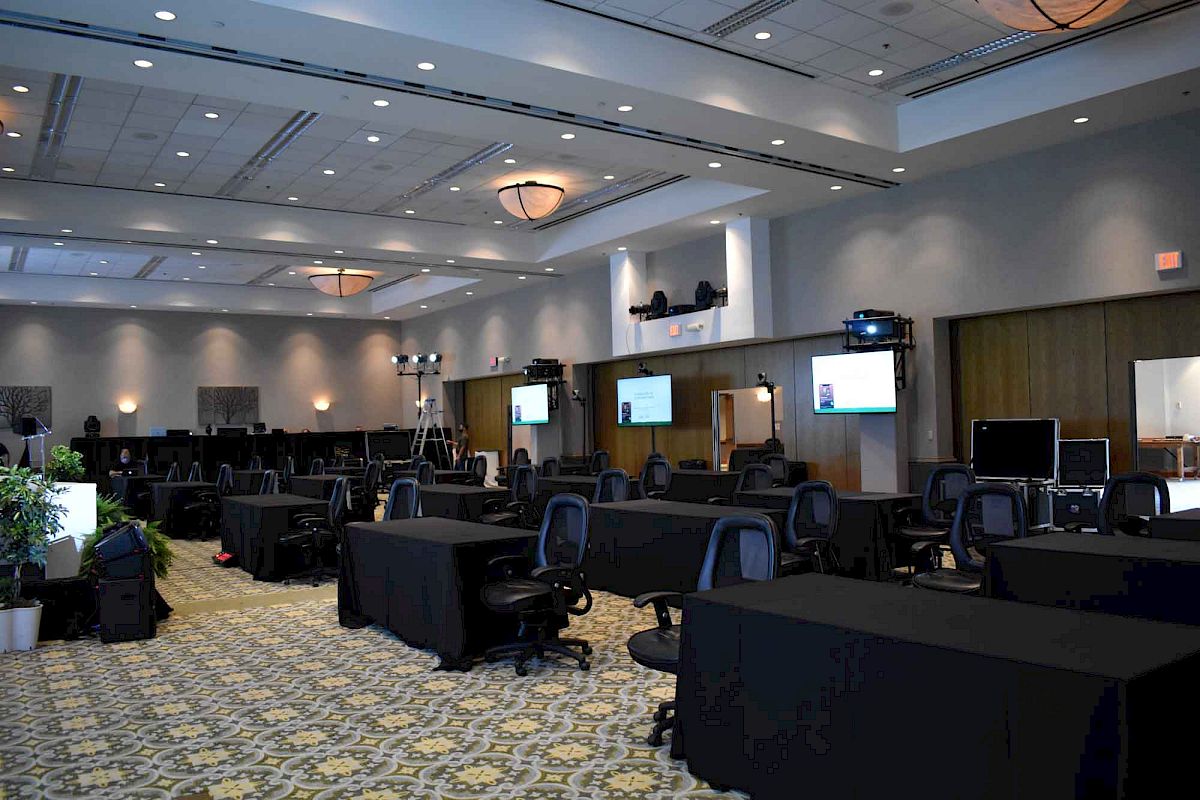 An empty conference room with rows of tables and chairs, television screens, lighting equipment, and a patterned carpet.