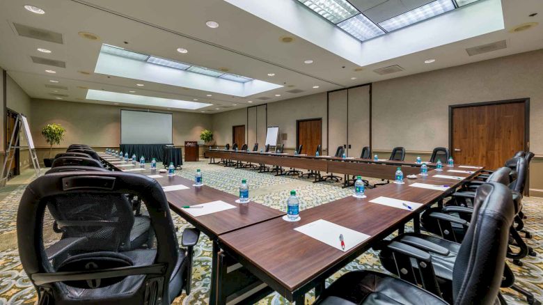 The image shows a conference room with a U-shaped table arrangement, chairs, notepads, pens, and bottled water set up for a meeting or presentation.