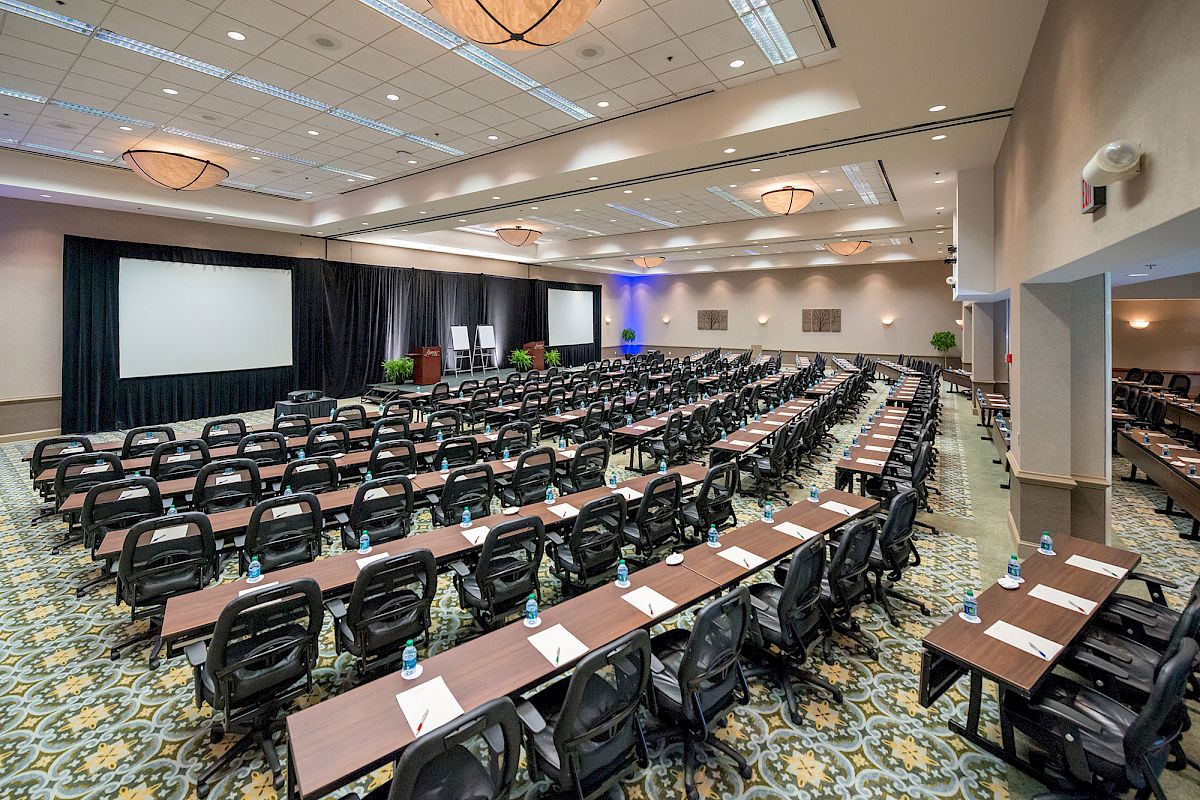 A large conference room set up with multiple rows of tables and chairs facing a stage with two screens and a podium, ready for an event or meeting.