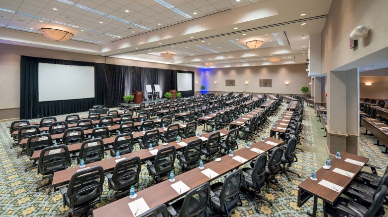 A large conference room set up with multiple rows of tables and chairs facing a stage with two screens and a podium, ready for an event or meeting.
