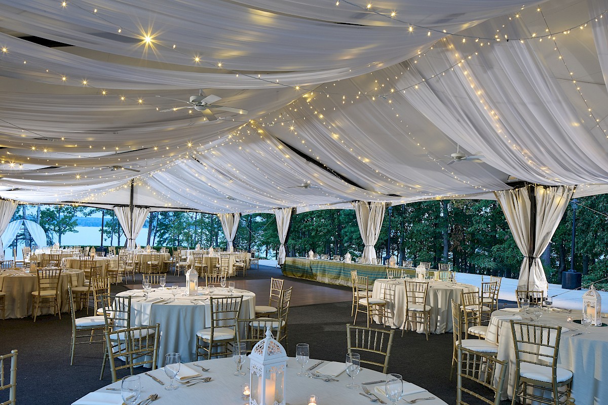 An elegant outdoor event space decorated with string lights and draped fabric, set with round tables and golden chairs for a celebration.